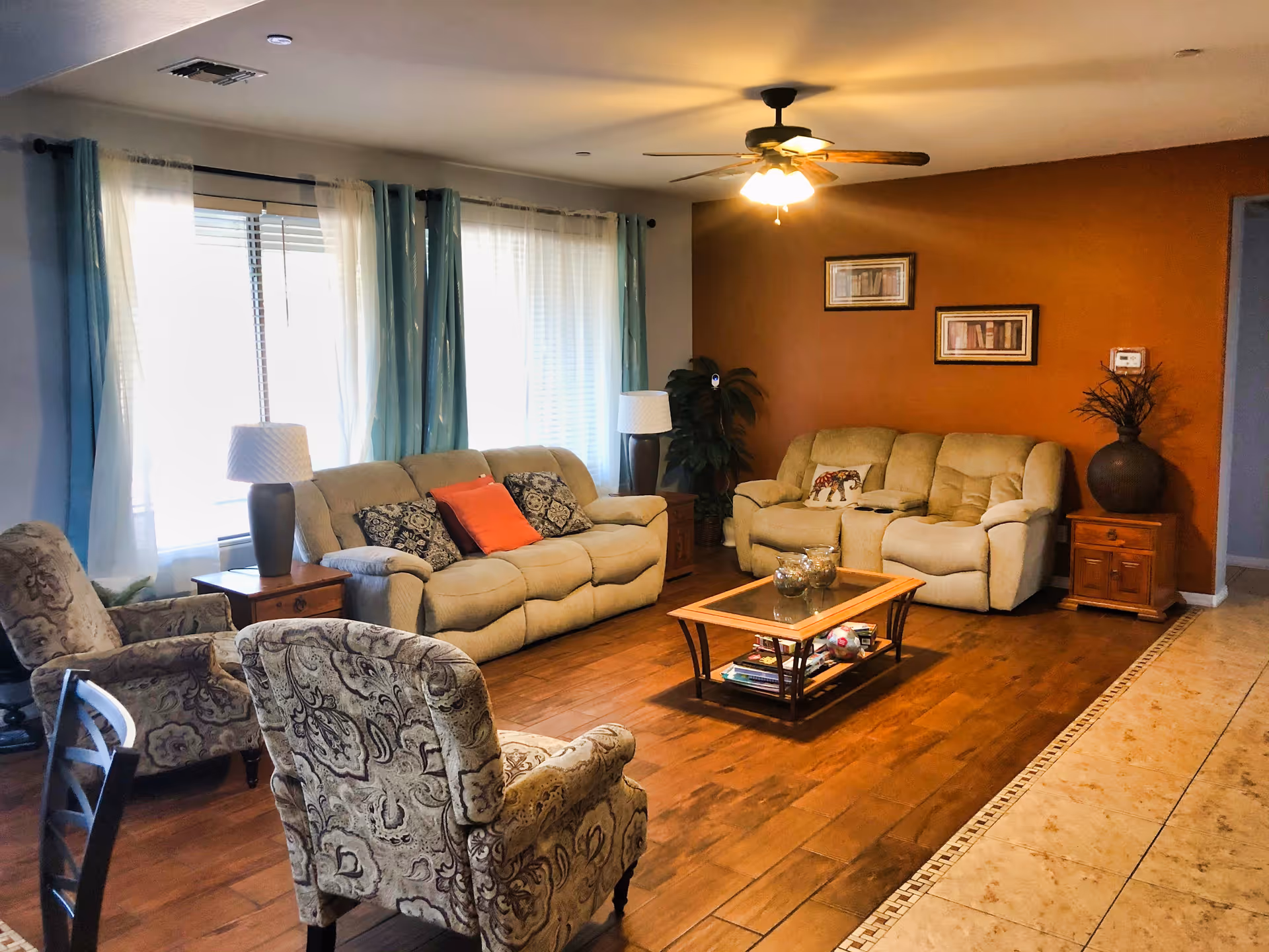 A cozy living room with two beige sofas, two patterned armchairs, a wooden coffee table with glass top, two side tables with lamps, and a ceiling fan with lights. The room has large windows with sheer and blue curtains, a wooden floor, and an orange accent wall with framed pictures and a large vase with decorative branches.