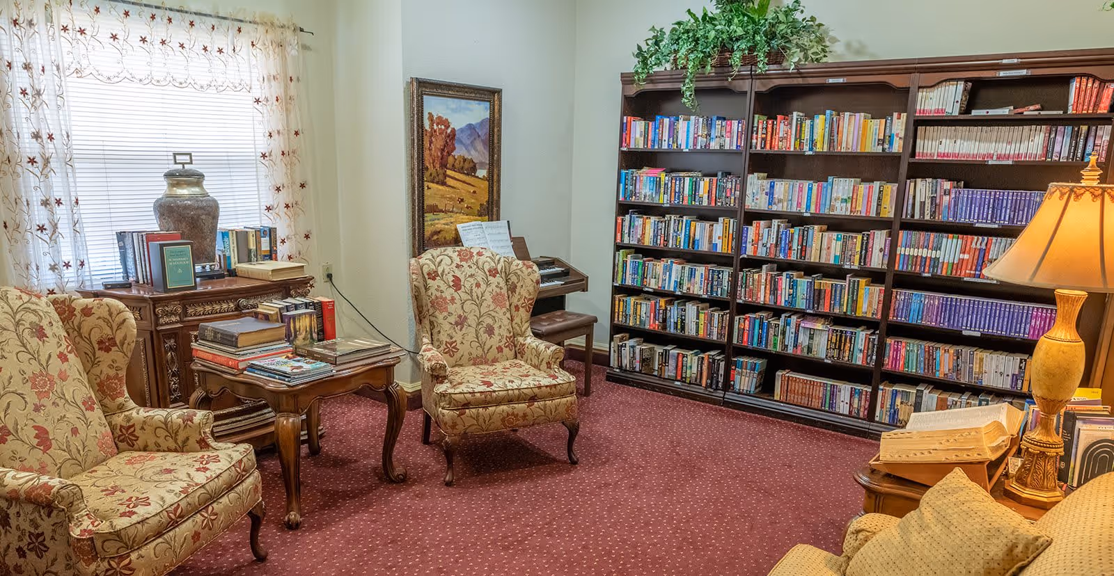 A cozy reading room with floral upholstered armchairs, wooden side tables stacked with books, a large bookshelf filled with books, a table lamp, a window with sheer floral curtains, and a framed landscape painting on the wall.
