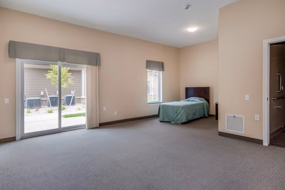A spacious bedroom with beige walls and carpeted floor featuring a single bed with a green bedspread near a window. There is a sliding glass door with vertical blinds leading outside, and an open doorway to a bathroom with grab bars visible.