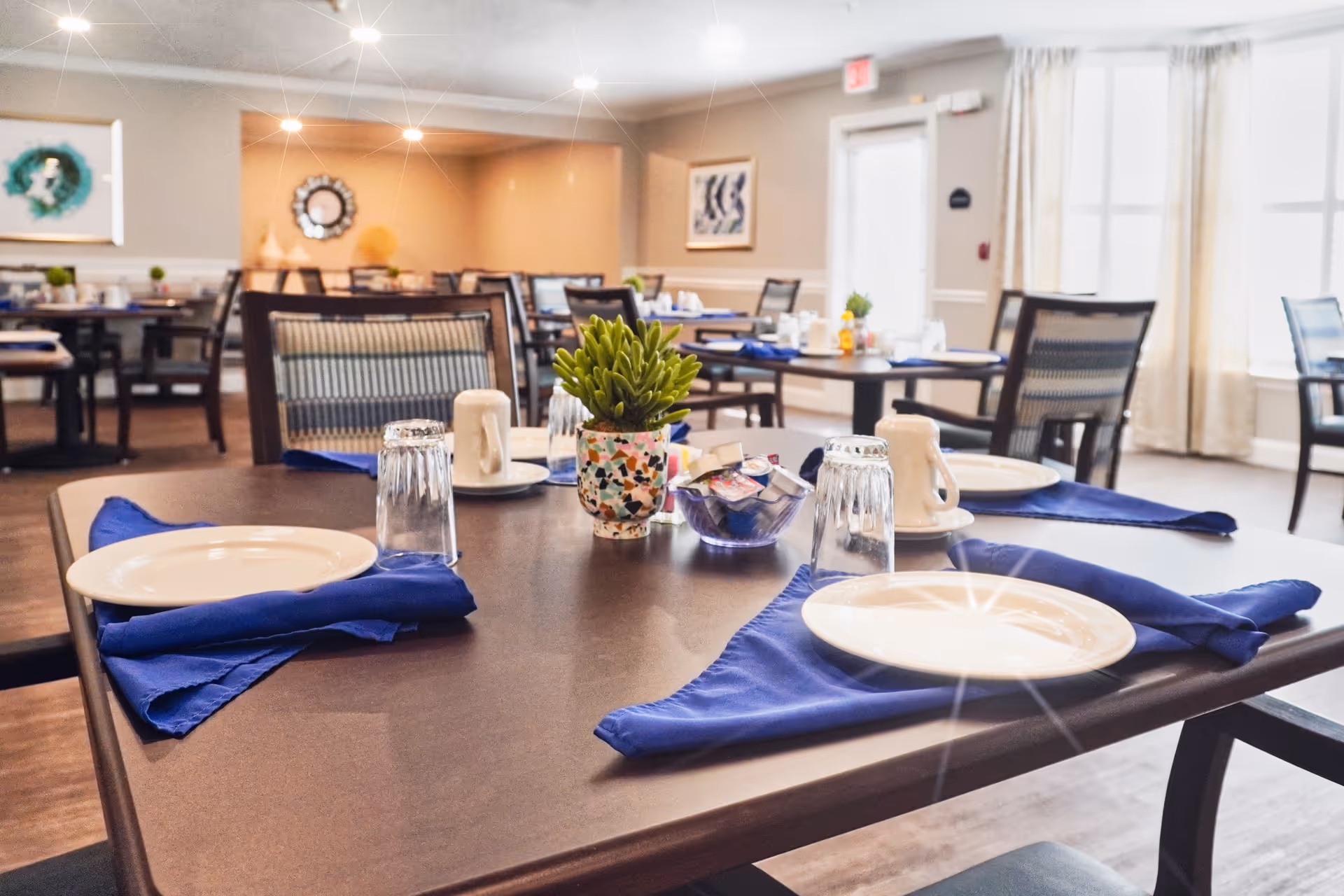 A dining room in a senior living facility with tables set for a meal. Each table has white plates, upside-down glasses, blue cloth napkins, and small decorative plants in colorful pots. The room is well-lit with natural light coming through large windows with curtains, and there are framed artworks on the walls.