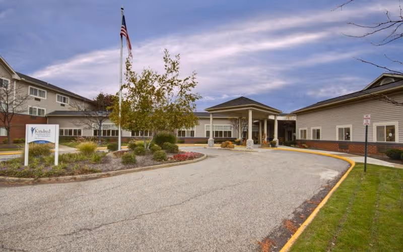 Front entrance and curved driveway of a senior living building with an American flag, landscaped island, and covered portico.