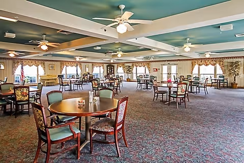 A spacious dining room with multiple round tables and chairs arranged throughout the room. The room has a patterned carpet, ceiling fans with lights, large windows with valances, and an American flag near the far wall. The room is well-lit with natural light coming through the windows.
