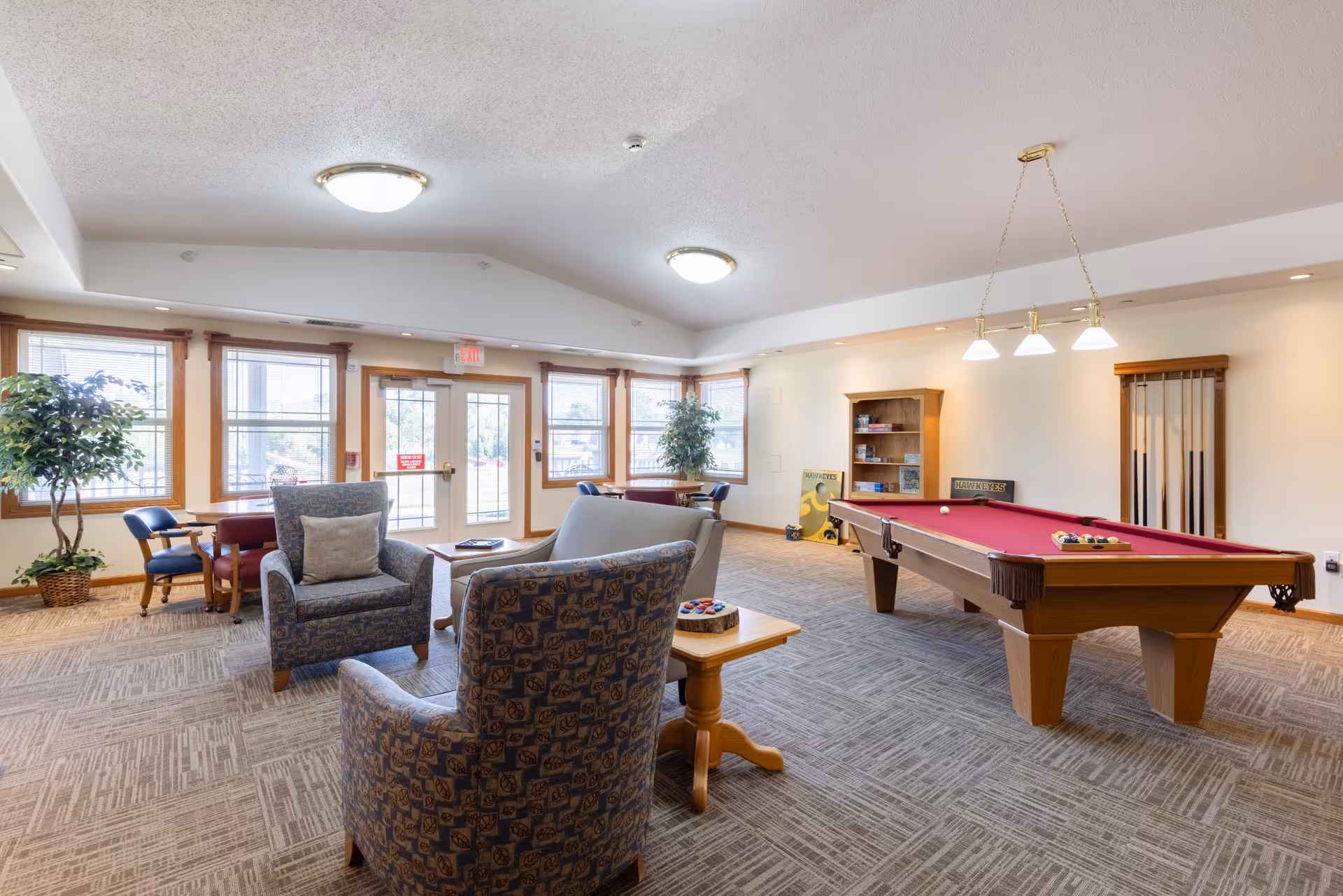 A spacious common room with a pool table featuring a red felt surface, surrounded by comfortable armchairs and small tables. The room has large windows allowing natural light to fill the space, potted plants, and a bookshelf in the corner. The ceiling has recessed lighting and a hanging light fixture above the pool table.