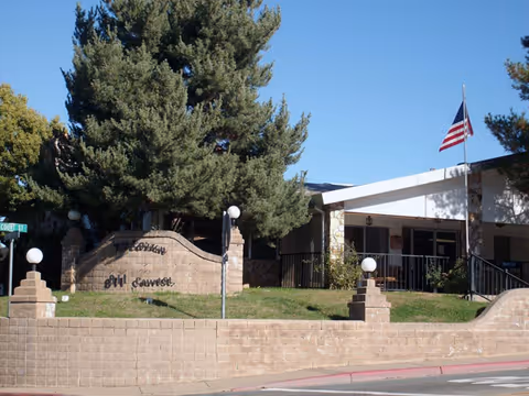 Front entrance of Kit Carson Nursing & Rehabilitation Center showing a brick sign and low wall, large trees, and an American flag.