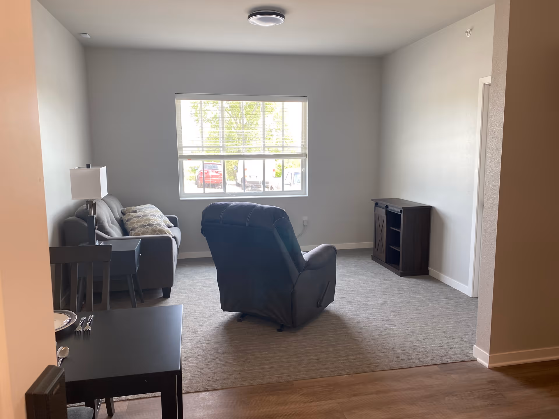 A small living room area with a gray sofa with patterned pillows, a dark recliner chair facing a window with blinds, a dark wooden side table with a lamp, a small dark wooden cabinet against the wall, and a dining table with chairs partially visible in the foreground.