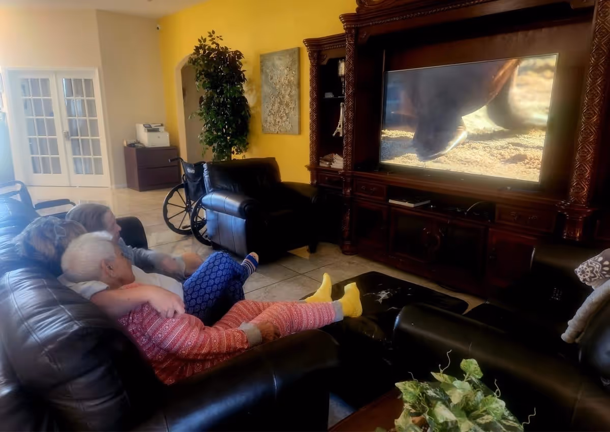 Two elderly women sitting on a black leather couch watching television in a cozy living room. One woman is wearing a red patterned outfit and yellow socks, while the other is in blue patterned pants. A wheelchair is visible near a black armchair, and there is a large wooden entertainment center holding the TV. The room has a yellow accent wall, a potted plant, and French doors in the background.