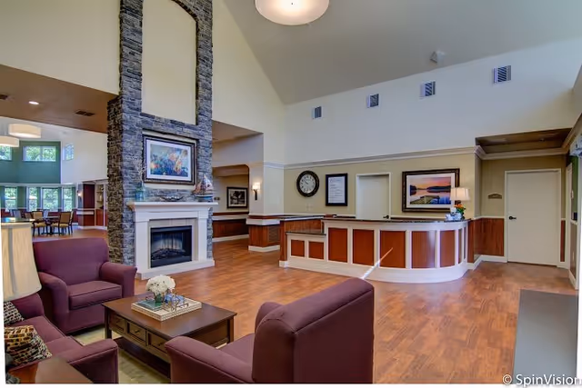 A spacious and well-lit senior living facility common area with wooden flooring, a stone fireplace, and comfortable purple armchairs around a wooden coffee table. The reception desk is visible in the background with framed artwork and a clock on the wall.
