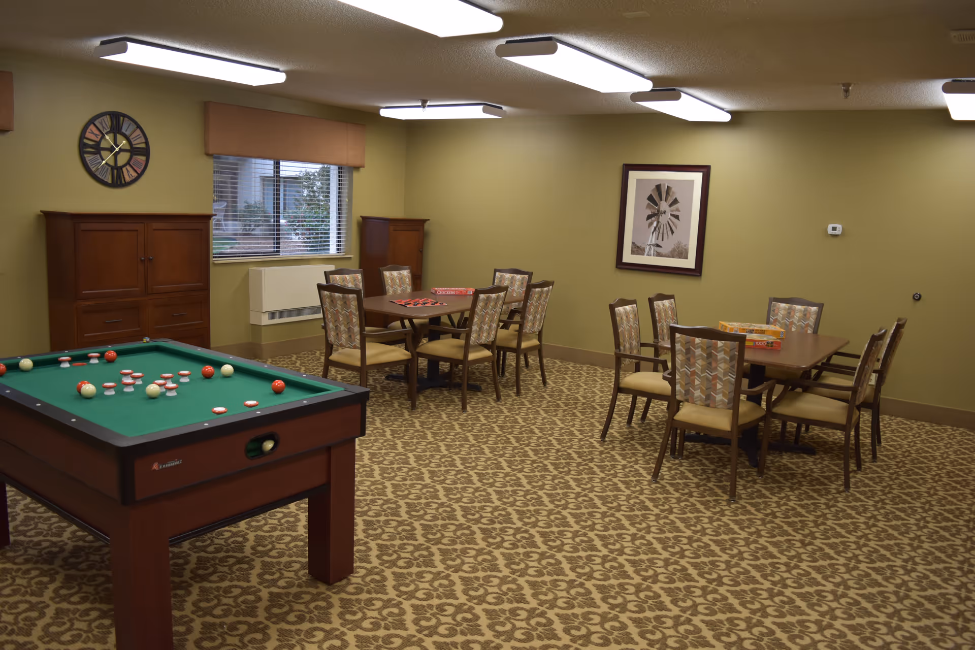 A game room with a pool table in the foreground and two tables with chairs in the background. The tables have board games and puzzles on them. The room has patterned carpet, green walls, a window with blinds, a clock on the wall, and a framed picture of a windmill.
