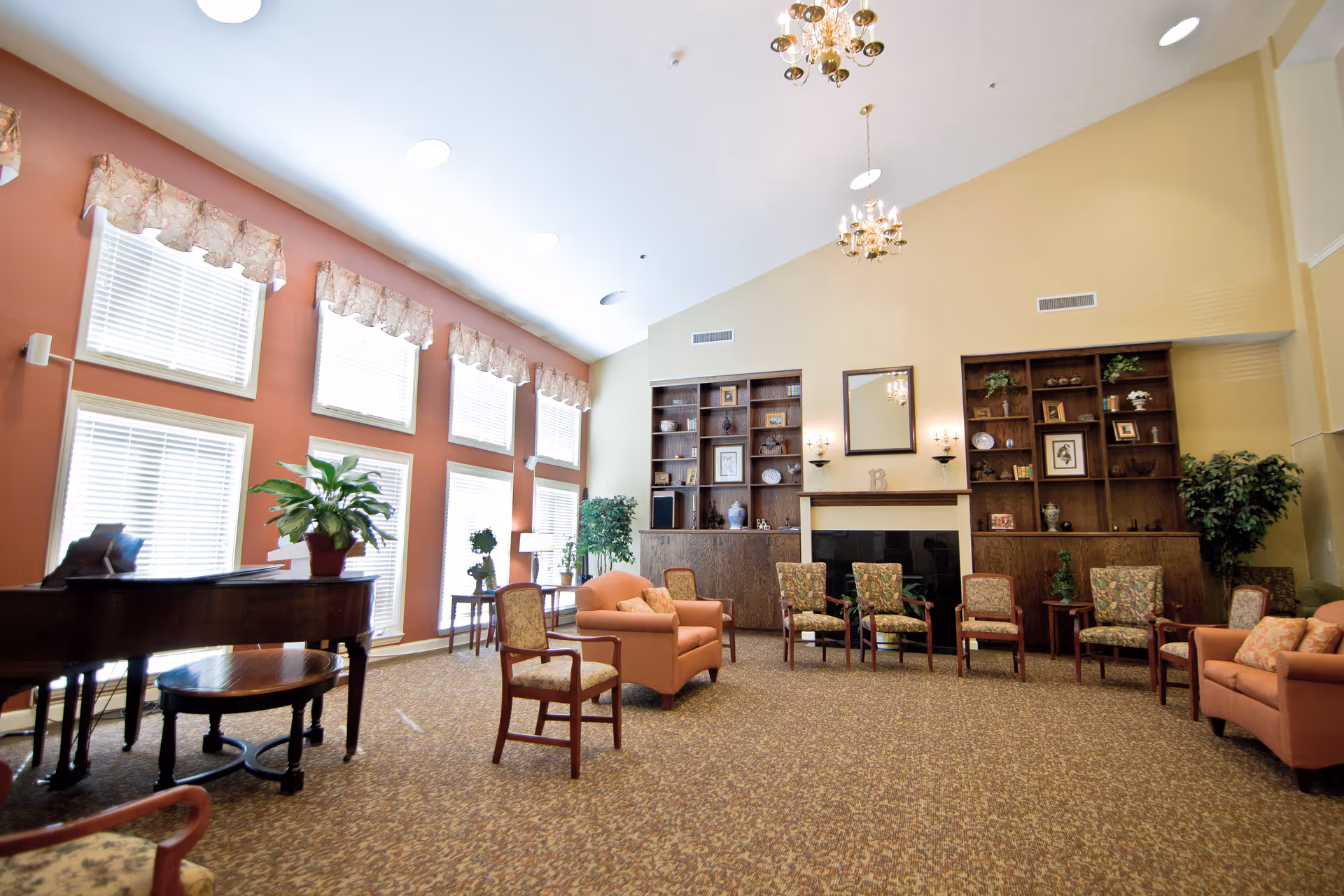 Spacious senior living common room with seating areas, bookshelves and a piano under large windows.
