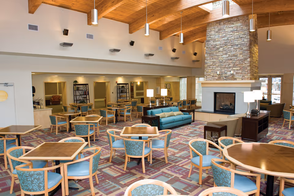 Spacious senior living common area with multiple wooden tables and chairs with blue cushions, a teal sofa, beige armchairs, a large stone fireplace, wooden ceiling beams, and modern pendant lights.