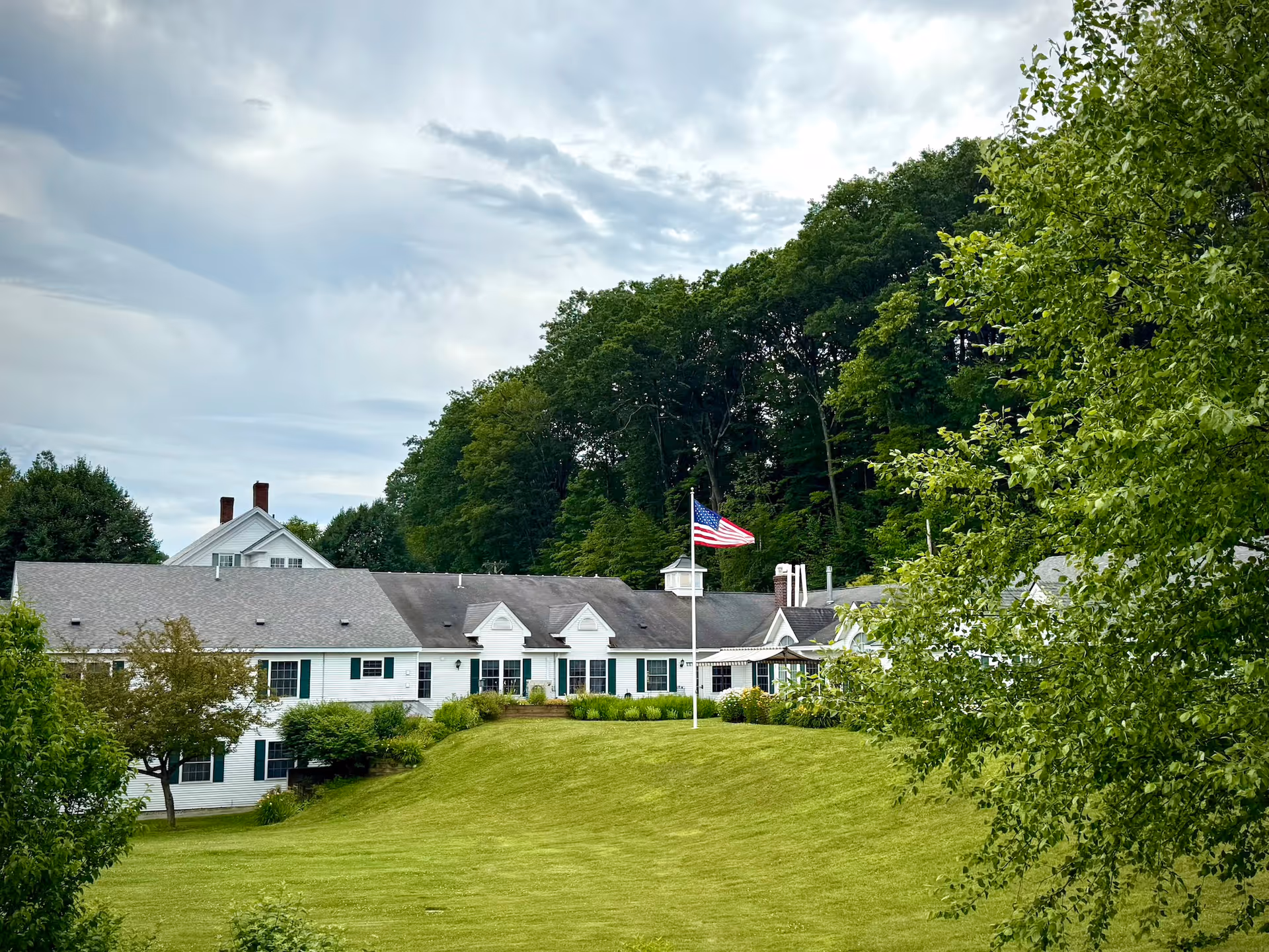 A large white building with green shutters situated on a grassy hill with an American flag on a flagpole in front. The building is surrounded by trees and under a cloudy sky.