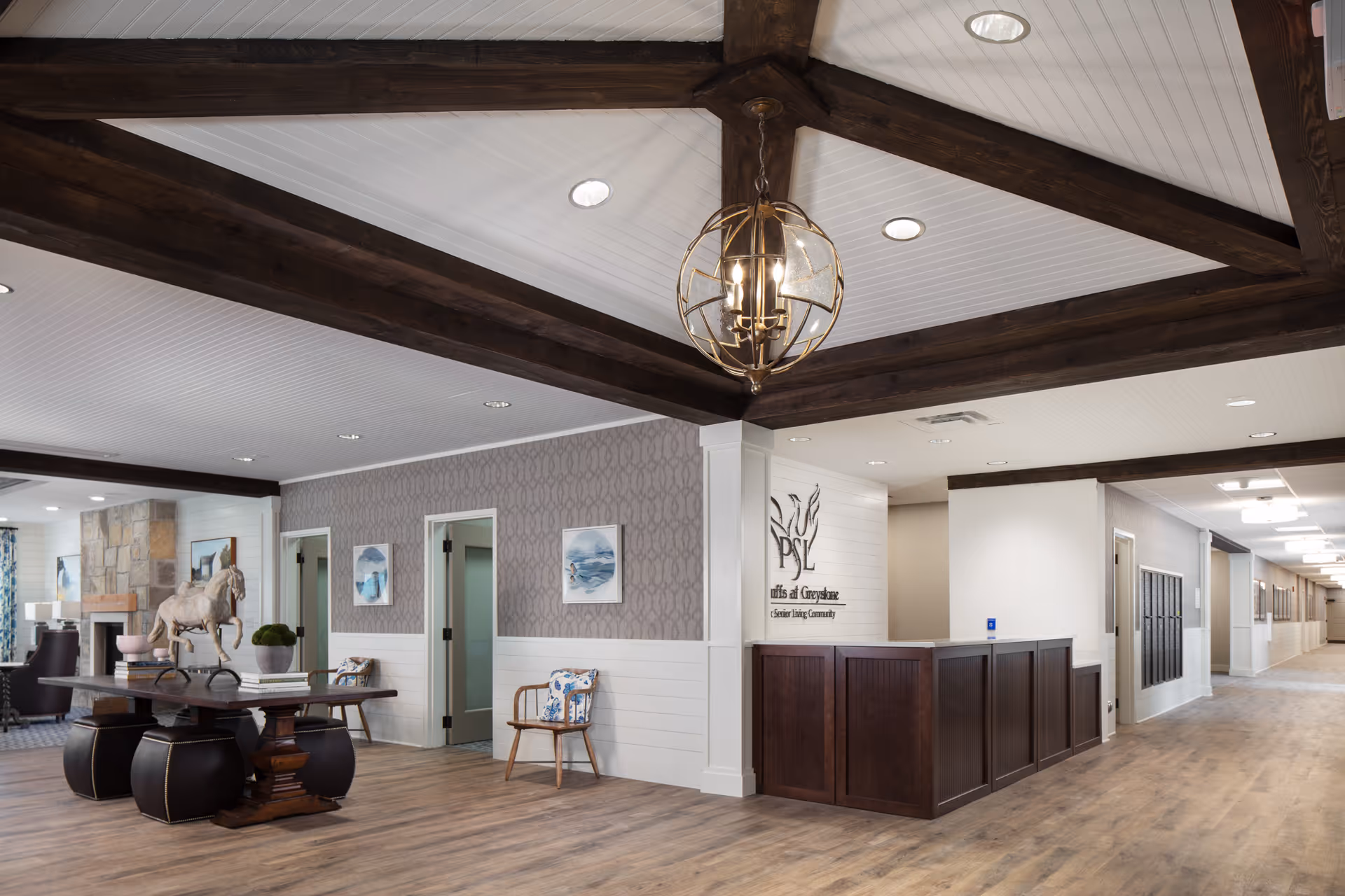 Interior view of Monark Grove Greystone senior living facility featuring a reception desk with dark wood paneling, a decorative chandelier hanging from a ceiling with exposed wooden beams, a hallway extending to the right, and a seating area with a wooden table, chairs, and a horse sculpture. The walls have patterned wallpaper and framed artwork.