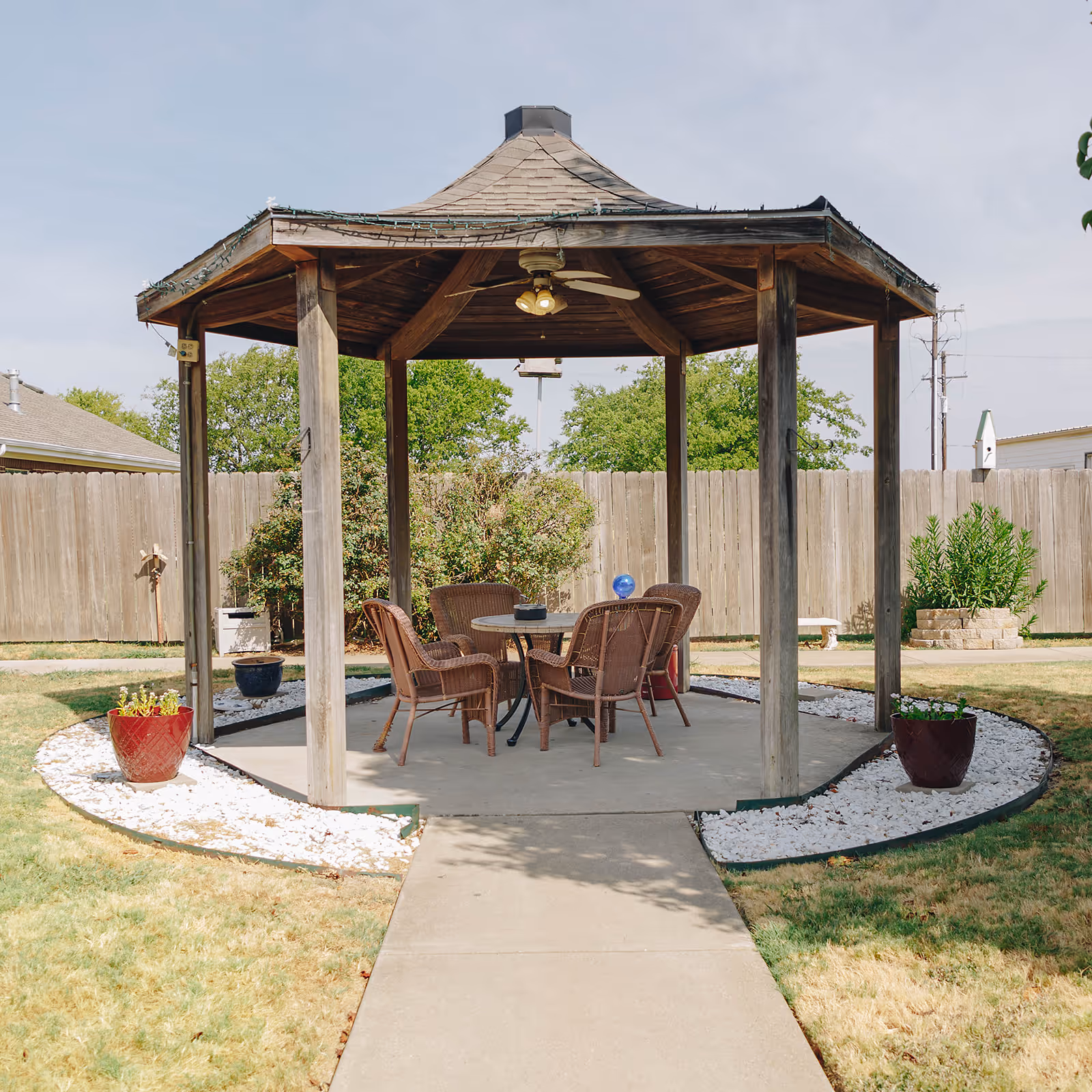 A wooden gazebo with a shingled roof and ceiling fan, situated on a concrete pad in a fenced backyard. Inside the gazebo are four wicker chairs around a round table. The surrounding area has grass, white decorative rocks, and potted plants.