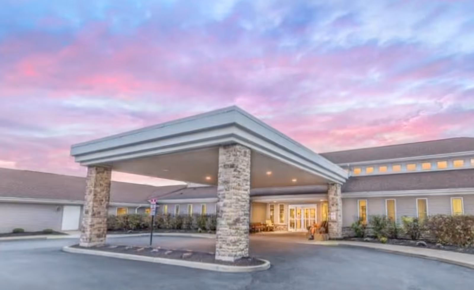 Exterior view of Brompton Heights of Williamsville building entrance with a covered drop-off area supported by stone pillars under a colorful sunset sky.