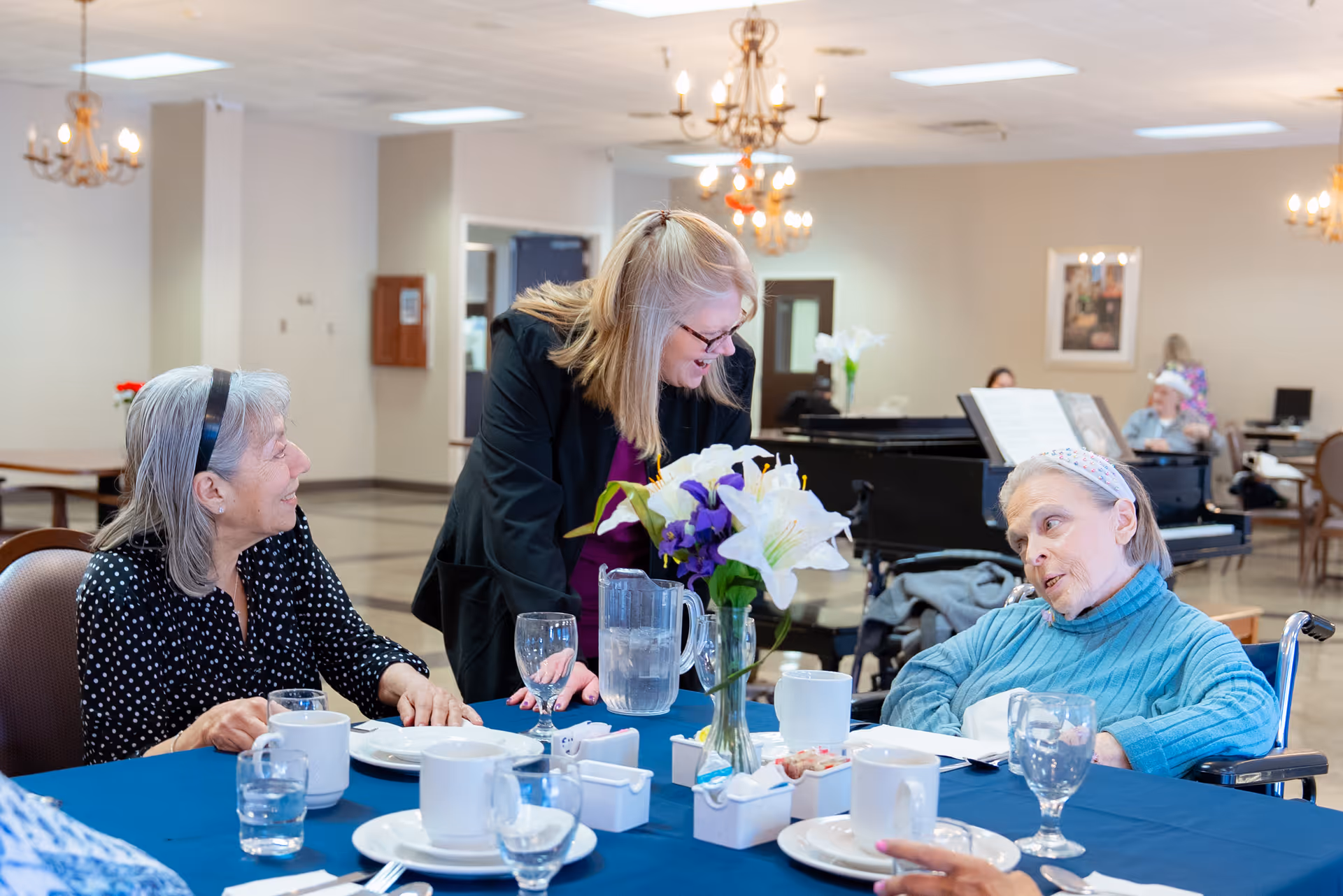 Three elderly women sitting around a dining table covered with a blue tablecloth, with cups, glasses, and plates. One woman in a wheelchair wearing a blue sweater is engaged in conversation with a standing woman who is smiling and leaning in. Another elderly woman with gray hair and a black polka dot shirt is also seated at the table. The background shows a piano and other people in a spacious, well-lit room with chandeliers.