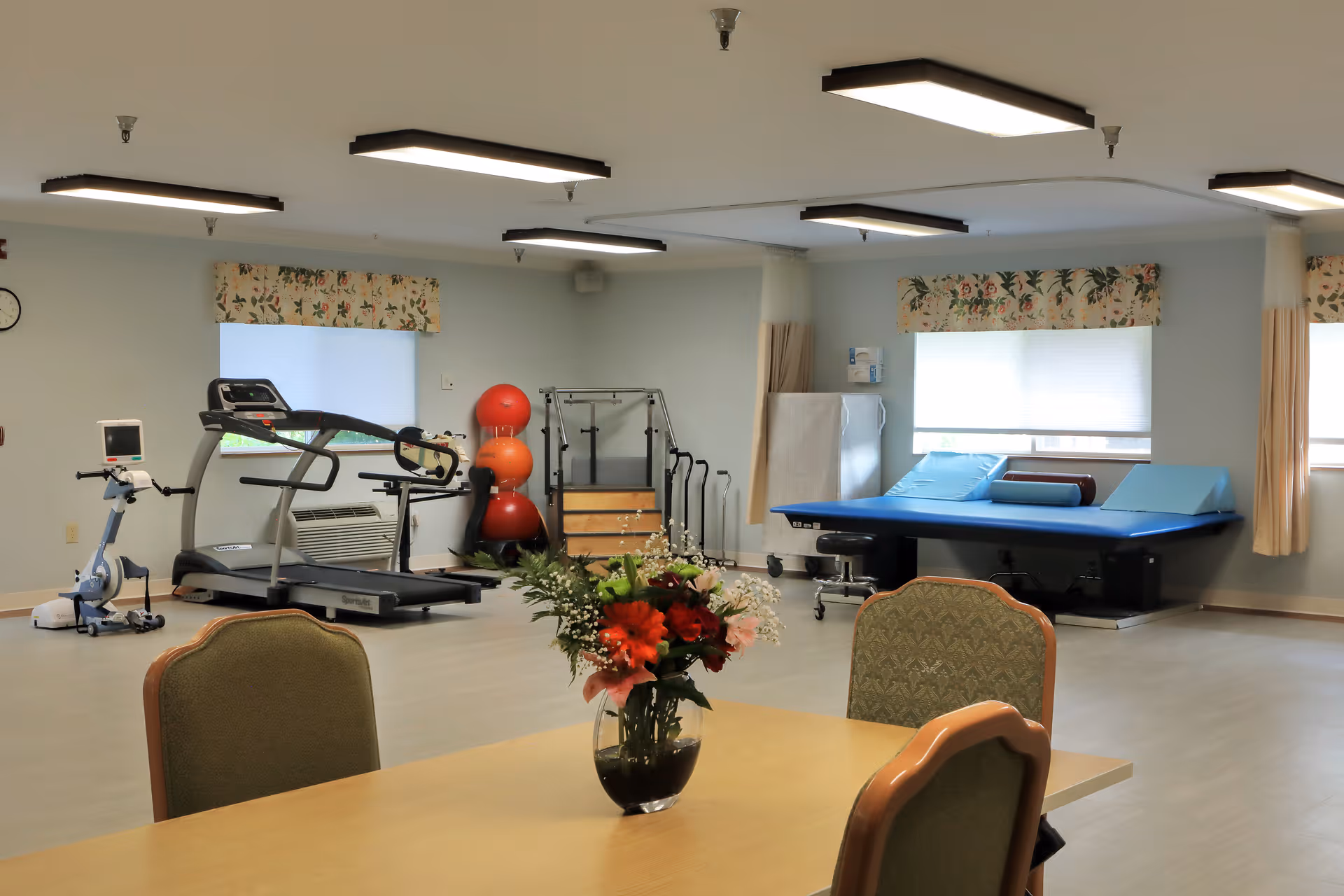 A bright interior therapy and exercise room with treadmills, exercise bike, therapy table, and a table with a vase of flowers.