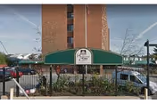 Exterior view of Highland Care Center showing a multi-story brick building behind a green awning with the facility's name and logo. There is a black metal fence and parked cars in front of the building.