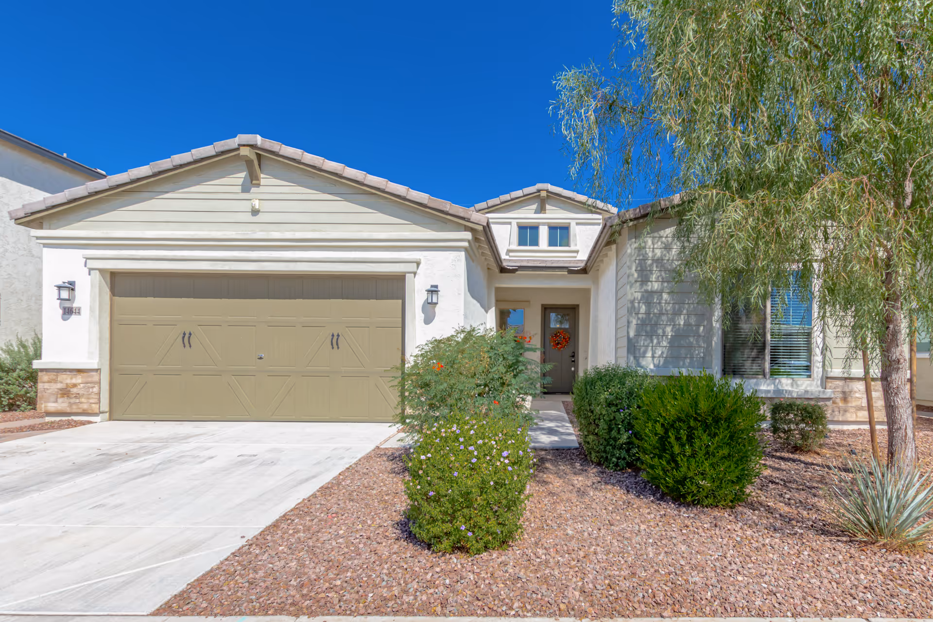 Front exterior view of a single-story house with a two-car garage, beige garage door, a pathway leading to the front door decorated with a wreath, and landscaping with bushes and a tree under a clear blue sky.