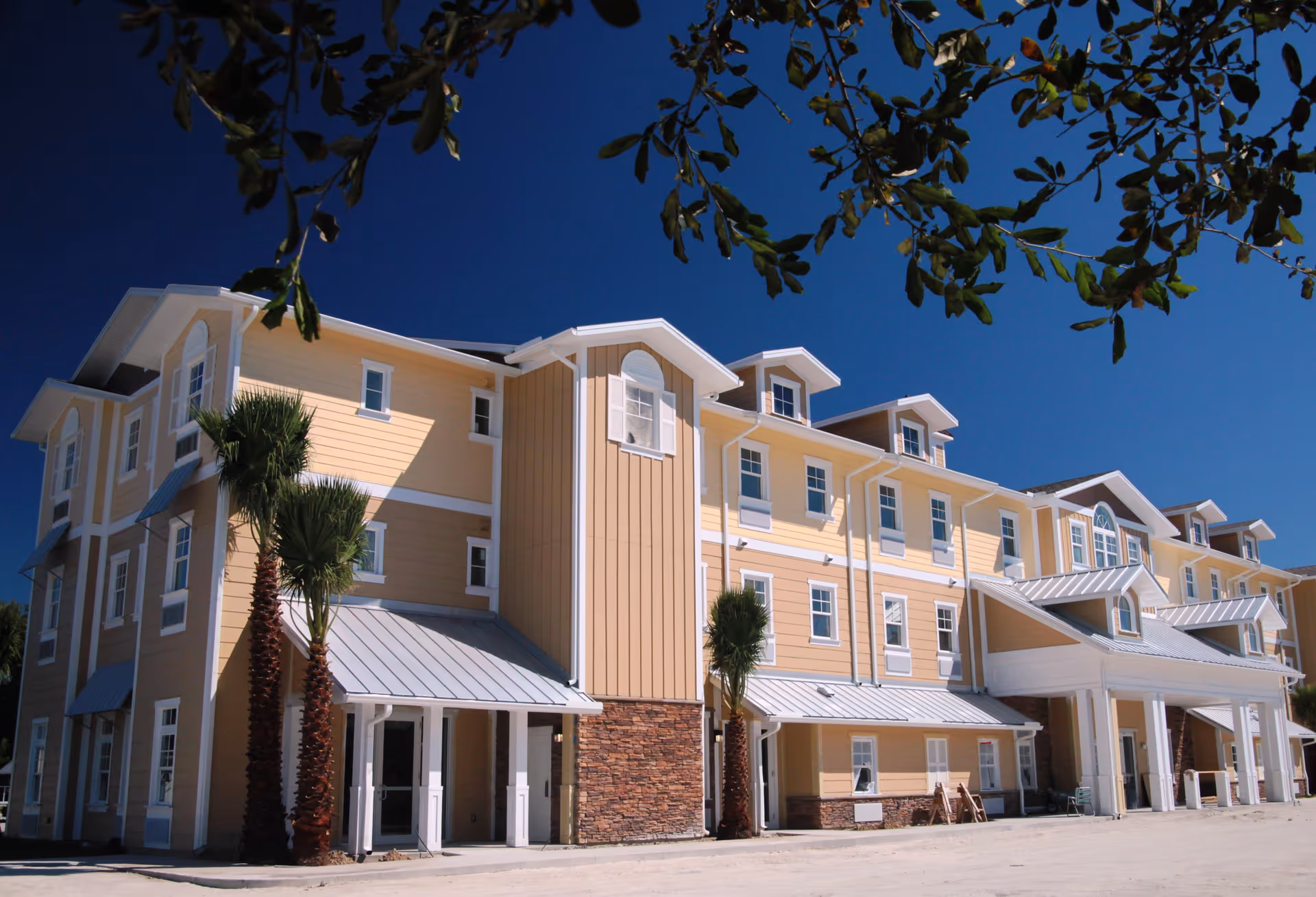 Exterior view of a large, multi-story senior living facility building with yellow siding, white trim, and stone accents. The building has multiple windows, metal awnings, and a covered entrance. Palm trees are planted near the building, and tree branches frame the top of the image against a clear blue sky.