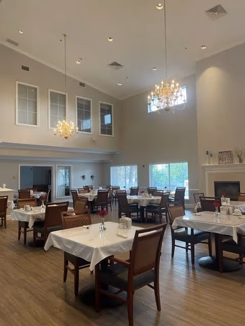 A spacious dining room with multiple tables covered in white tablecloths, each set with chairs and small vases containing red flowers. The room features high ceilings with two chandeliers, large windows letting in natural light, and a fireplace on the right side. The flooring is wood, and the walls are painted in neutral tones.