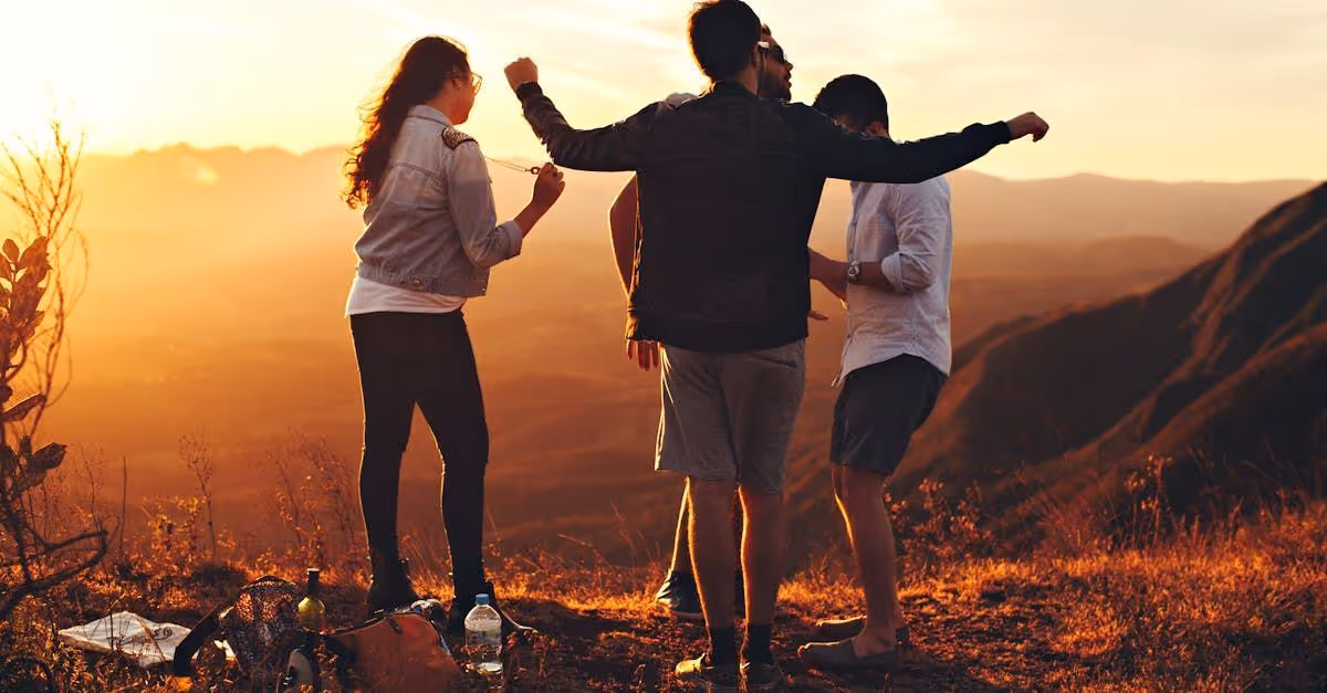 Four people stand on a hilltop at sunset overlooking a mountainous landscape with backpacks and a water bottle on the ground.