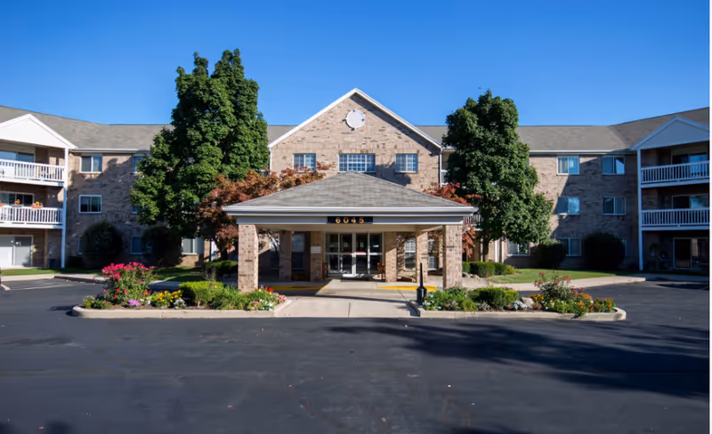 Three-story brick senior living building with a covered entrance, landscaped flowerbeds and a paved driveway under a clear blue sky.