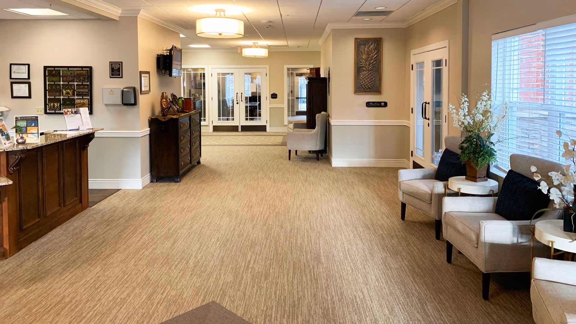 A clean and well-lit reception area in a senior living facility with a wooden reception desk on the left, beige carpeted floor, comfortable beige armchairs with black cushions on the right, small round tables with flower arrangements, and double glass doors at the far end.