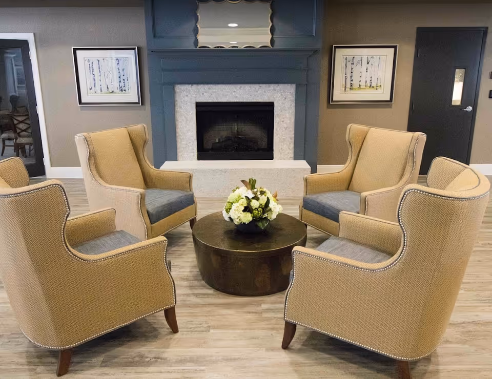 A cozy seating area with four beige armchairs arranged in a circle around a round wooden coffee table with a floral centerpiece. Behind the seating area is a fireplace with a blue mantel and two framed pictures on the wall. The room has light wood flooring and neutral-colored walls.
