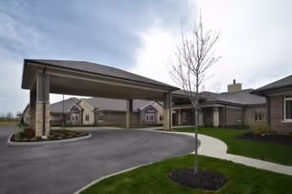 Exterior view of The Ganzhorn Suites showing a covered driveway entrance with a circular driveway, landscaped grass areas, and a leafless tree in the foreground under a cloudy sky.