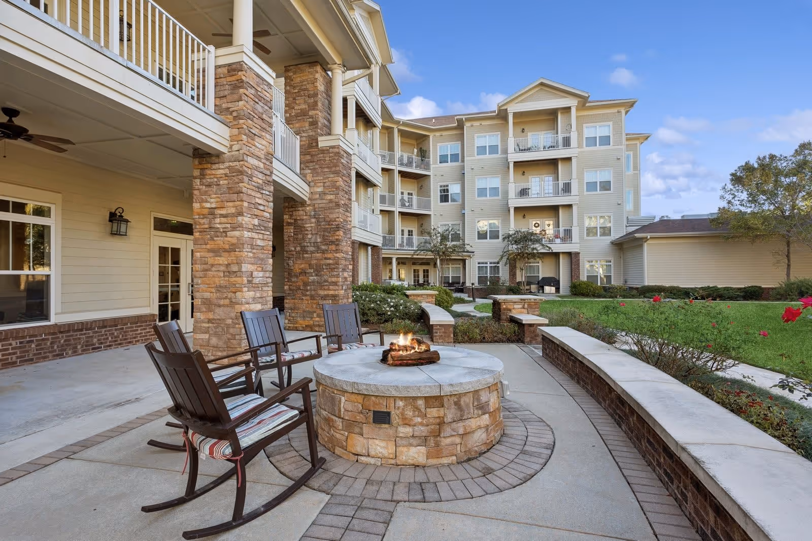 Outdoor seating area at a senior living facility with a round stone fire pit surrounded by four wooden rocking chairs with cushions. The area is paved with concrete and brick accents, and there is a multi-story building with balconies and stone pillars in the background. Green grass, bushes, and a few red flowers are visible on the right side under a partly cloudy sky.