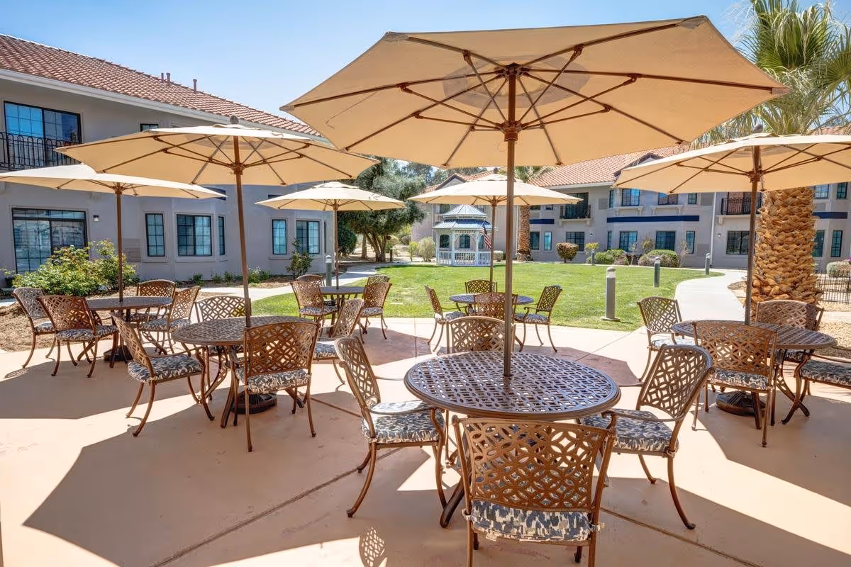 Outdoor patio area with multiple round metal tables and chairs with patterned cushions, each table shaded by large beige umbrellas. The patio is surrounded by a green lawn, palm trees, and two-story residential buildings with windows and balconies in the background. A white gazebo is visible in the distance.