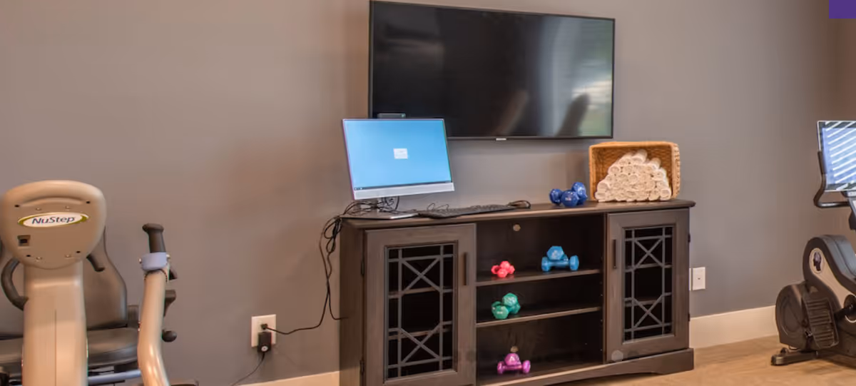 A fitness room with exercise equipment including a NuStep machine and another cardio machine. A black cabinet holds colorful dumbbells and a basket of rolled white towels. A computer monitor and a large flat-screen TV are mounted on the wall above the cabinet.