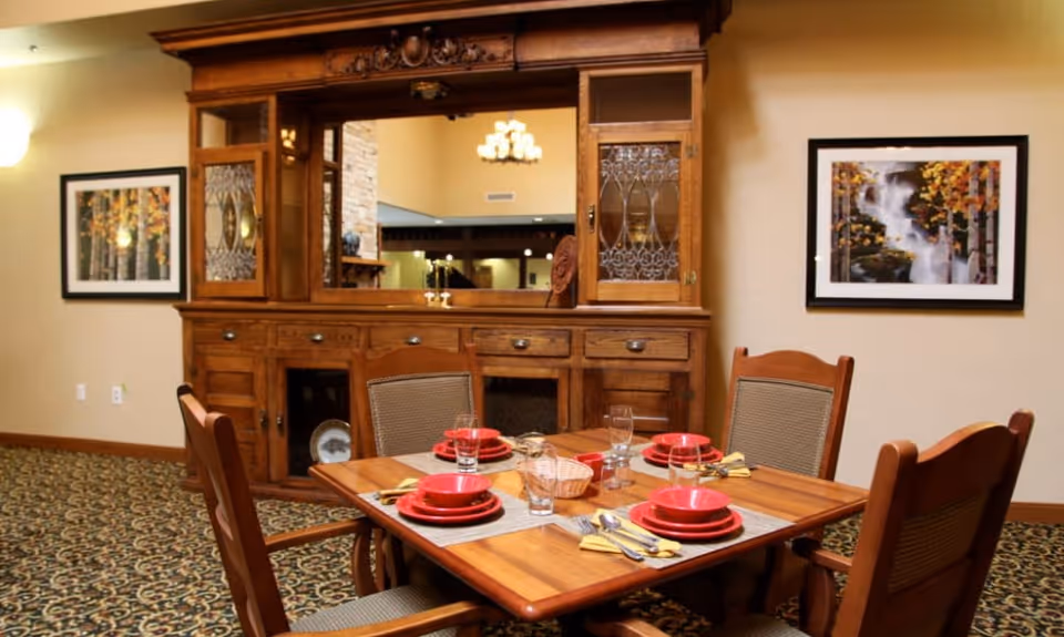 A dining area with a wooden table set for four people with red bowls, plates, glasses, and yellow napkins. The table is surrounded by four wooden chairs with cushioned seats. Behind the table is a large wooden cabinet with glass doors and a mirror. The walls are decorated with framed pictures of nature scenes, and the floor is carpeted with a patterned design.