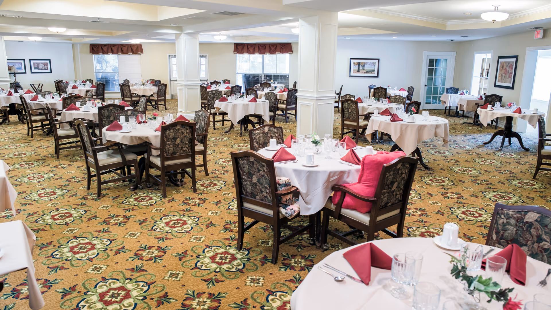 Spacious dining room with multiple round tables set with white linens and red napkins, floral carpet and upholstered chairs.