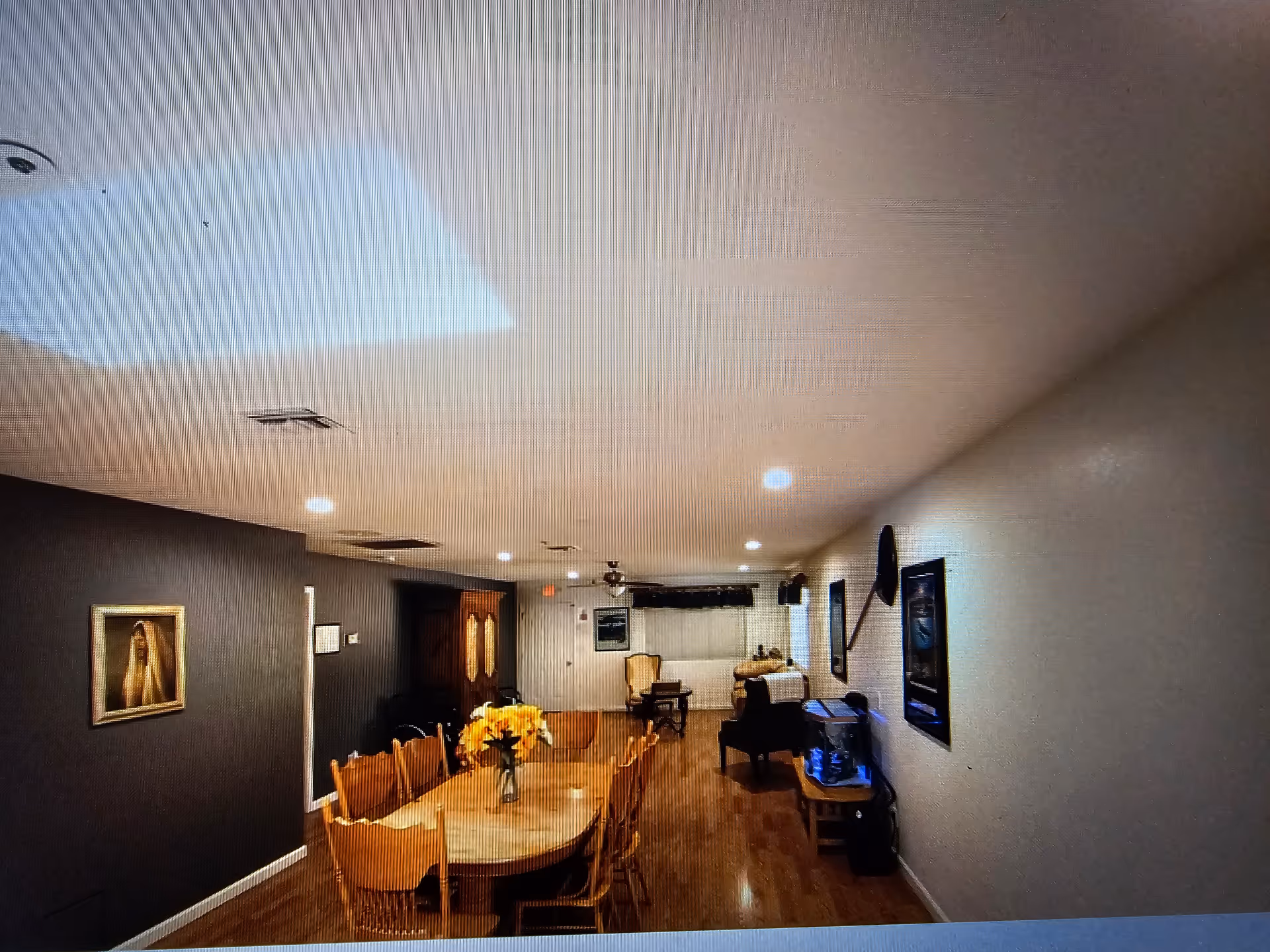 A long room with a wooden dining table and chairs in the foreground, a vase with yellow flowers on the table. The room has wooden flooring, recessed ceiling lights, and a ceiling fan. There are framed pictures on the walls, a fish tank on a small table, and armchairs towards the back of the room.