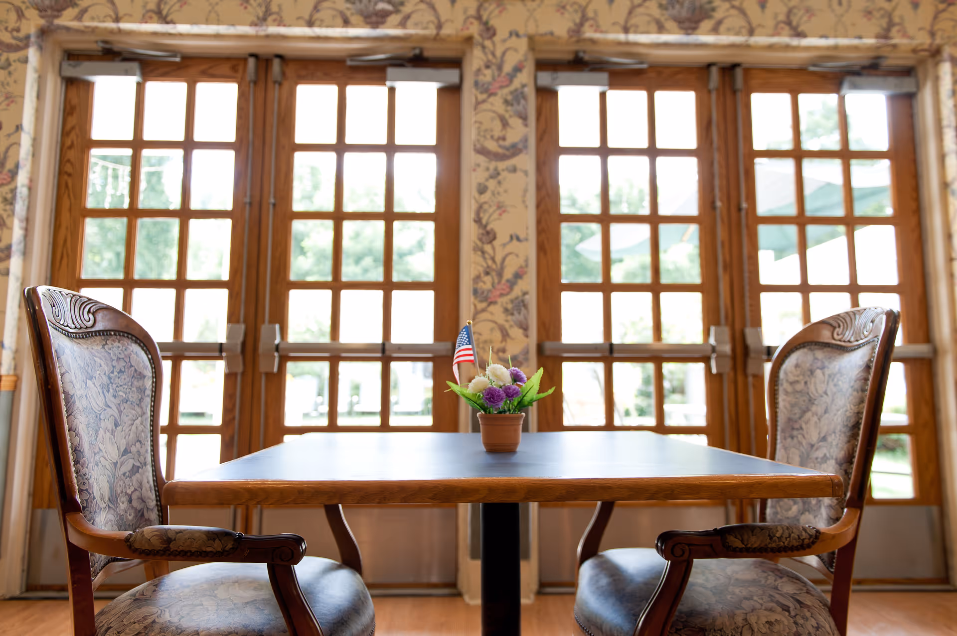 A wooden table with a small flower pot containing purple and white flowers and a miniature American flag in the center, flanked by two upholstered chairs with floral patterns. Behind the table are large wooden-framed glass doors letting in natural light and showing greenery outside.