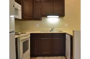 Small kitchen area with dark wooden cabinets, a white stove with oven, a microwave above the stove, a sink with a faucet, and a dishwasher. The walls are light-colored, and there is under-cabinet lighting illuminating the countertop.