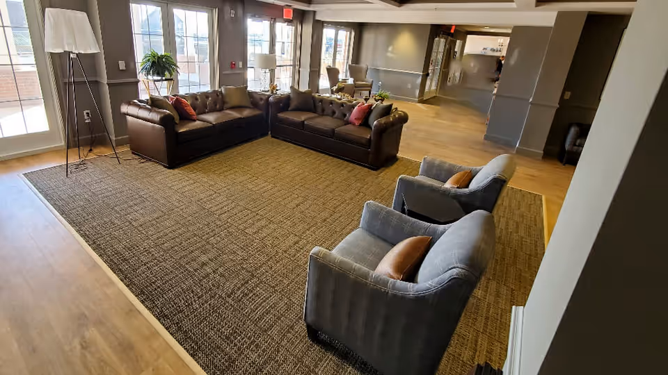 Spacious common lounge with leather sofas and upholstered armchairs arranged on a large area rug near windows.