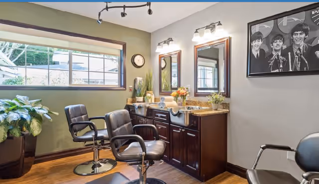 Interior view of a salon area with two black salon chairs in front of a wooden vanity with two sinks and mirrors. There are plants on the vanity and a large potted plant near the window. A black and white framed photo of The Beatles hangs on the wall.