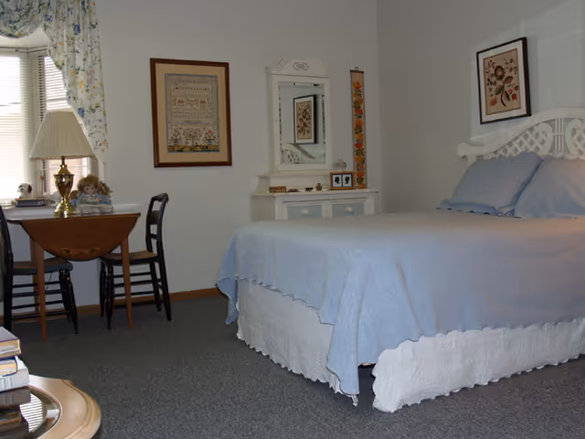A bedroom with a neatly made bed covered in light blue bedding and white bed skirt. There is a white headboard with a framed floral picture above it. To the left of the bed is a white dresser with a mirror and some small decorative items. A small wooden table with two chairs is placed near a window with floral curtains. A lamp and two dolls are on the table. The walls are light-colored and there are framed pictures hanging on them.