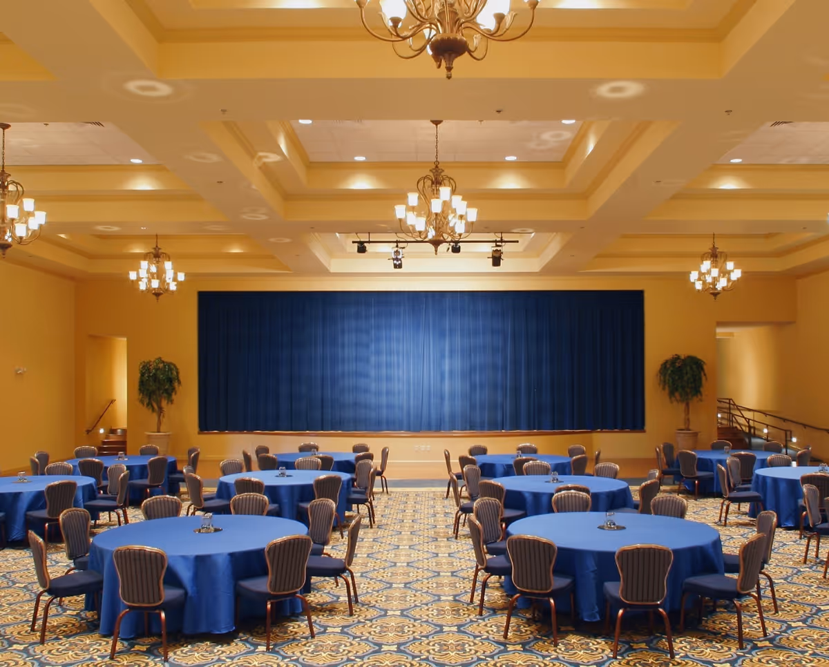 A large banquet hall with round tables covered in blue tablecloths and surrounded by chairs. The room features a patterned carpet, yellow walls, multiple chandeliers hanging from a coffered ceiling, and a stage with a closed blue curtain at the far end. There are potted plants on either side of the stage.
