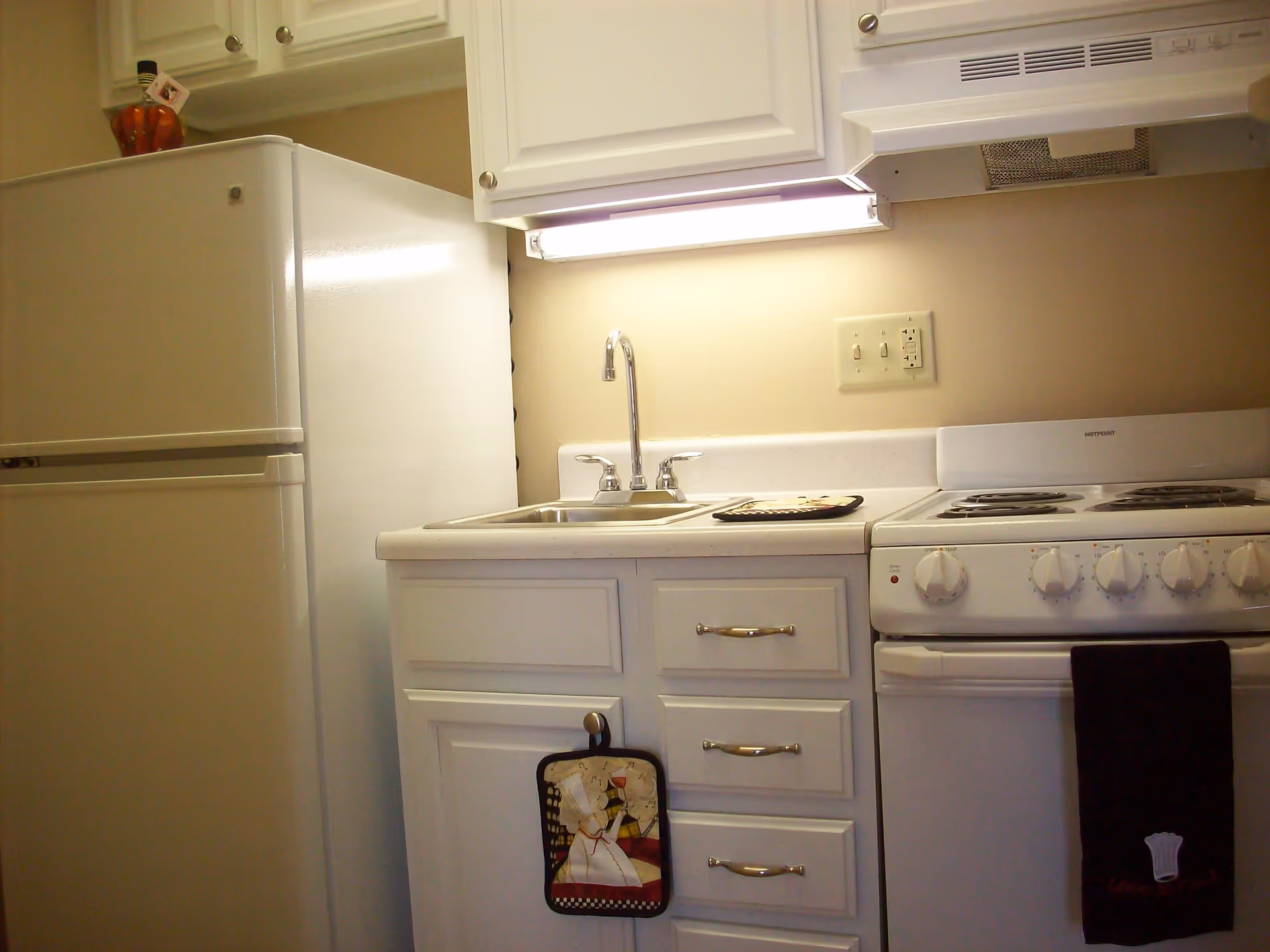 A small kitchen area with a white refrigerator, white cabinets, a stainless steel sink with a faucet, a white electric stove with four burners, and a black towel hanging on the oven door. There is a decorative oven mitt hanging on a cabinet handle and a light fixture under the cabinet illuminating the countertop.