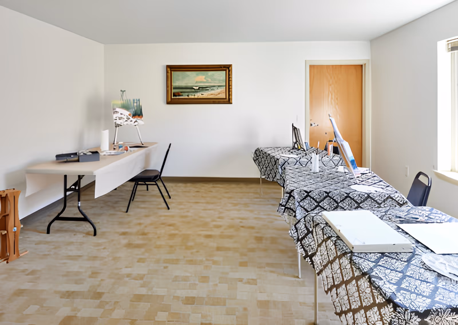 A simple room with two tables covered with patterned cloths and art supplies, including canvases and paintbrushes. There are two chairs, a painting of a beach scene on the wall, a closed wooden door, and a window letting in natural light.