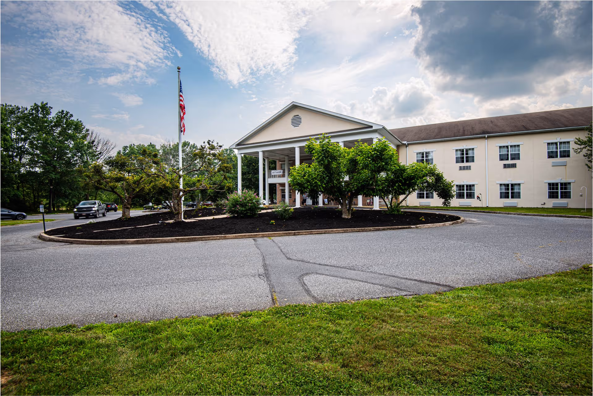 Front exterior of a nursing and rehabilitation center with a columned entrance, circular driveway, flagpole, and landscaped trees under a cloudy sky.