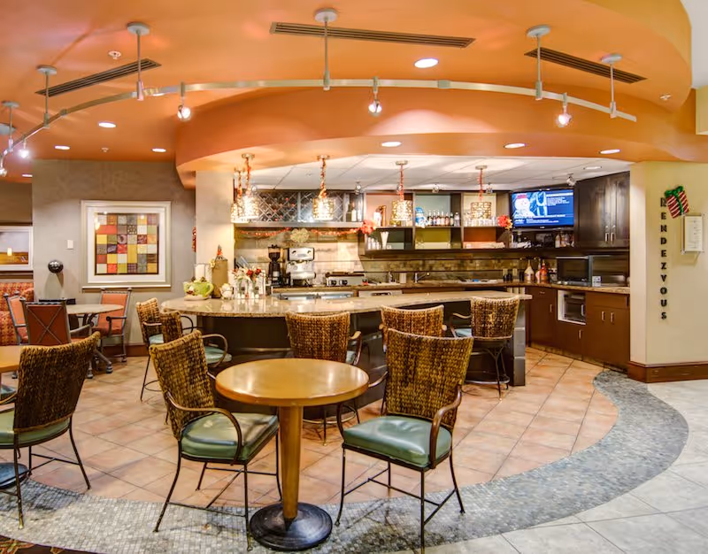 Cozy dining and lounge area with round tables, wicker chairs, and a central bar counter under warm lighting.