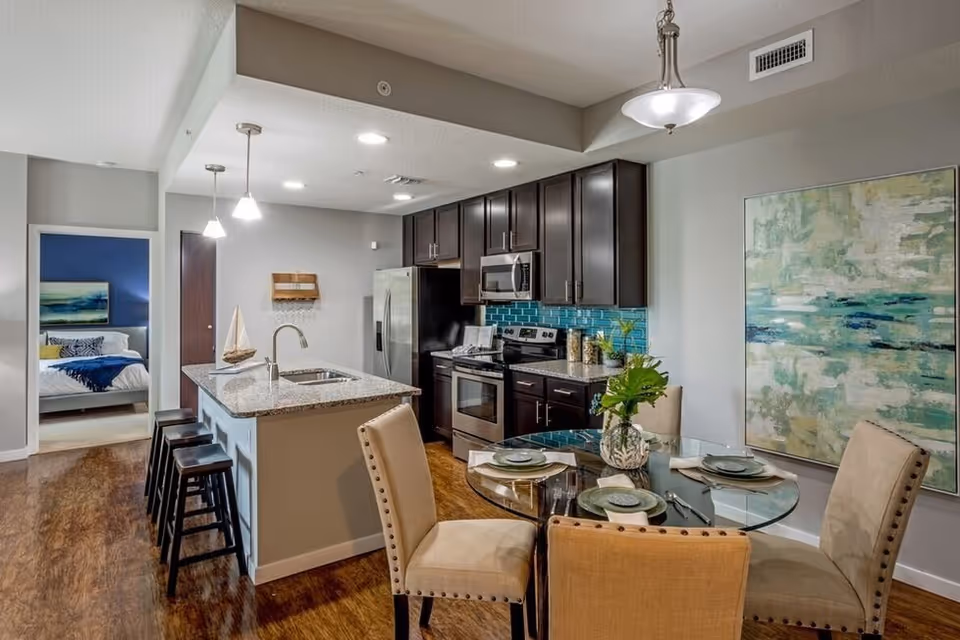 Modern kitchen and dining area with dark wood cabinets, stainless steel appliances, a kitchen island with bar stools, and a round glass dining table set with plates and napkins. A bedroom with a bed and artwork is visible through an open doorway.
