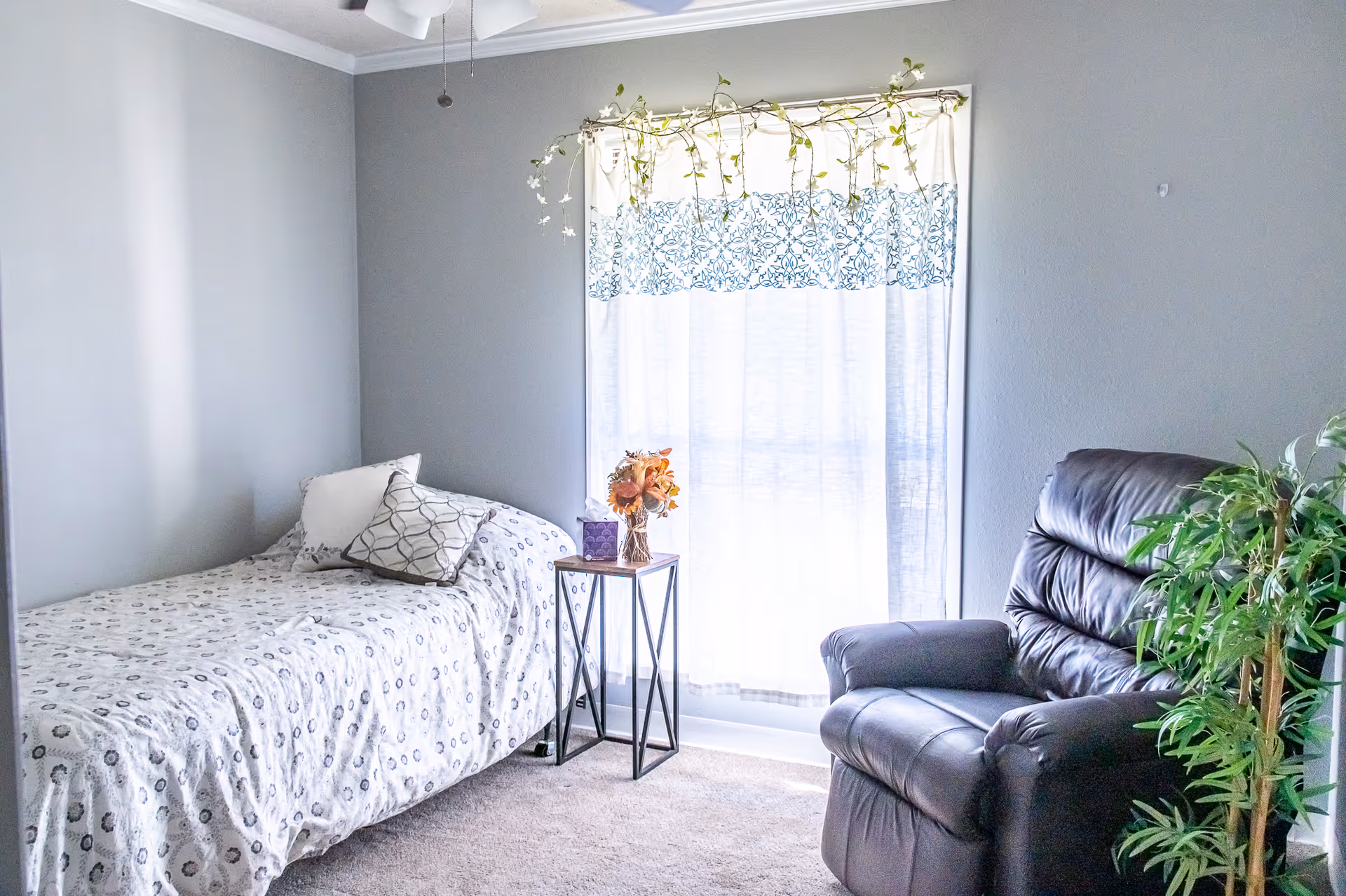 A small, cozy bedroom with a single bed covered in a white and gray patterned bedspread, two pillows, a small side table with a vase of flowers and a tissue box, a large window with white and blue patterned curtains, a black leather recliner chair, and a green potted plant in the corner.