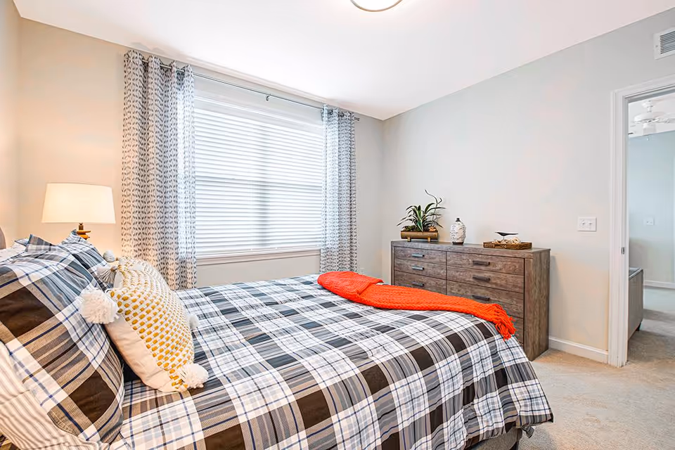 A bright bedroom with a large window covered by patterned curtains. The bed has a plaid bedspread with a decorative pillow and an orange throw blanket. A wooden dresser with plants and decorative items is against the wall, and an open doorway leads to another room.