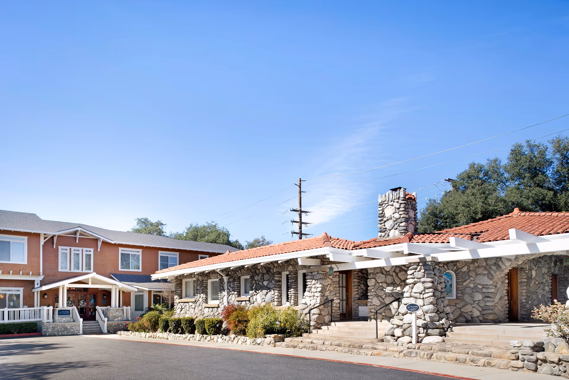 Exterior view of Ivy Park at Claremont facility showing a stone building with a red tiled roof and a chimney, alongside a two-story building with brown walls and white trim under a clear blue sky.
