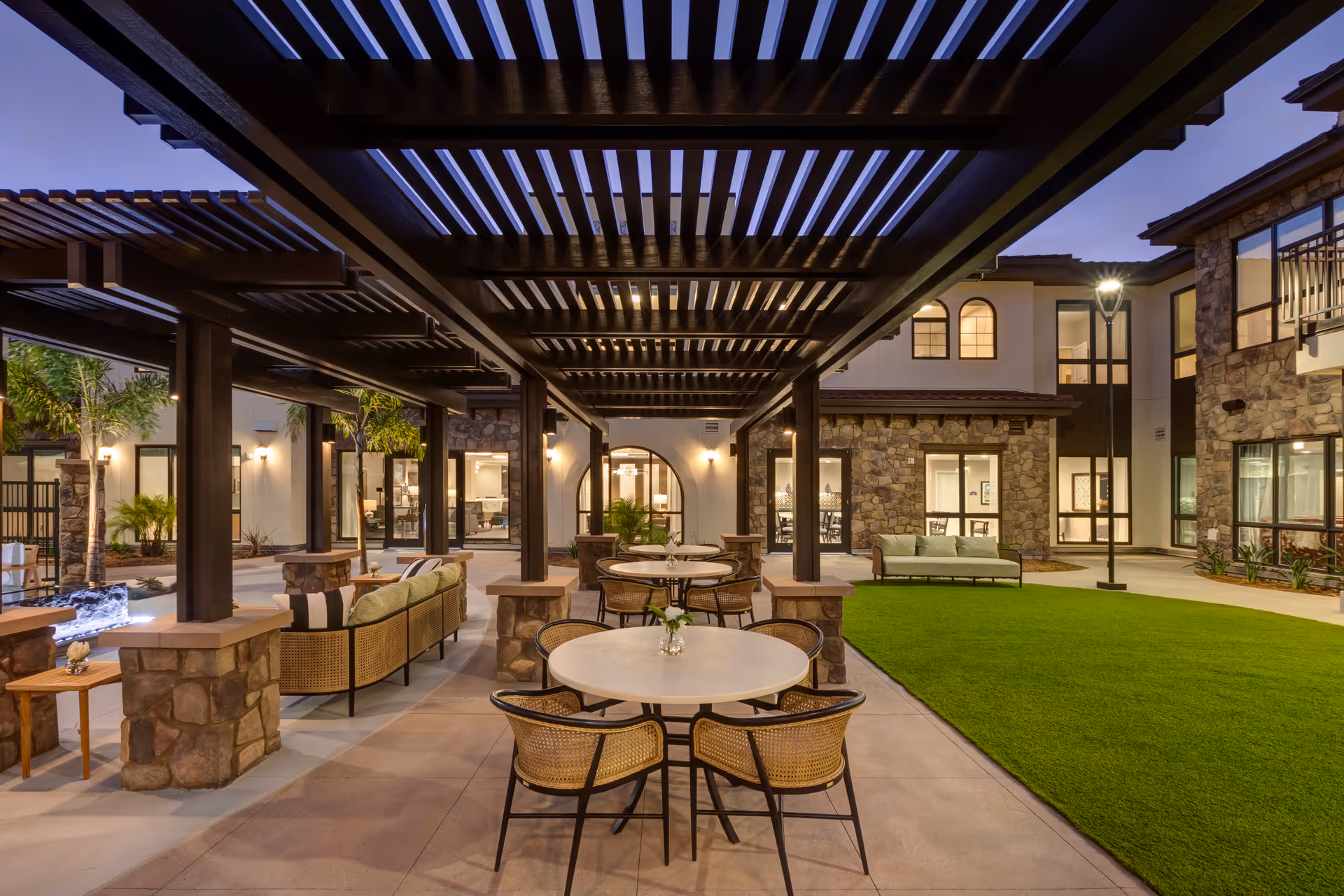Outdoor courtyard area at dusk with stone pillars supporting a wooden pergola. Several round tables with wicker chairs and cushioned sofas are arranged on a paved patio next to a well-maintained grassy area. The building surrounding the courtyard has stone and stucco walls with large windows and warm interior lighting.